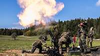 Fünf Soldaten hocken und stehen neben einem Panzermörser im Gras. Mit einem Feuerblitz verlässt die Granate das Rohr.