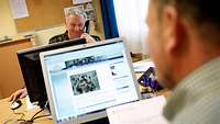 Ein Soldat sitzt an seinem Schreibtisch und telefoniert, während im Vordergrund ein Kollege an seinem PC arbeitet.