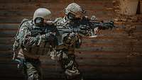 Zwei Soldaten zielen mit einem Gewehr vor einer braunen Holzwand.