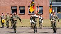 Drei Soldaten marschieren nebeneinander. Der Soldat in der Mitte trägt die Truppenfahne.