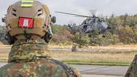 Soldat im Feldanzug mit Gefechtshelm steht vor einem tief schwebenden Transporthubschrauber. 