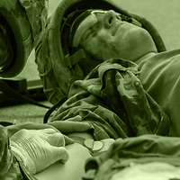 A medical orderly applies an intravenous catheter to the arm of an injured fellow soldier lying on the ground.