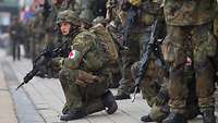 An armed medical orderly kneels in position on a pavement. Numerous other soldiers stand close by.