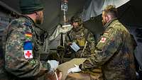 In a medical tent, two medics attend to a casualty while another soldier, standing next to them, conveys information.