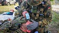 A soldier wearing a protective respiratory mask uses water to rinse the eye of another soldier lying on the ground.