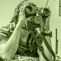 A soldier uses binoculars to observe from within a vehicle.