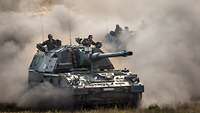 Two armoured self-propelled howitzers drive across a training area, throwing up large clouds of dust.