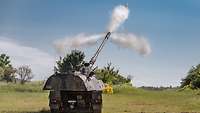 A Panzerhaubitze 2000 on a training area with sparse vegetation fires off a round, emitting a smoke plume.