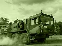 Army logistics personnel provide the force Raising a cloud of dust, a heavy equipment transporter carries a Fuchs armoured transport vehicle.