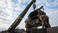 A crane keeps the powerpack of a Leopard 2 main battle tank suspended in mid-air. Two repairmen inspect the powerpack.