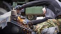 A serviceman lies on his side and uses an abrasive cutting machine on a truck’s frame, producing a shower of sparks.