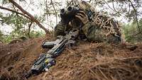 A soldier lies close to a small earth mound and observes through a telescopic sight.