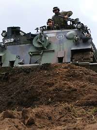 Ein Panzer mit einem Räumschild schiebt Erdreich vor sich her. Aus der Luke schauen zwei Soldaten.
