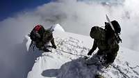 Two soldiers rappel on a snow-covered rock ledge in high mountains; one of them acts as belayer.