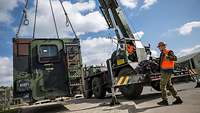 A crane with a container as suspended load. It is guided into place by a soldier pulling a rope. 