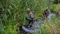 Zwei grün getarnte Soldaten laufen durch das Wasser entlang eines Baches.