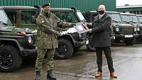 Ein Soldat und ein Zivilist halten einen großen symbolischen Zündschlüssel. Im Hintergrund stehen fünf Geländewagen.