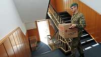 Ein Soldat, in Uniform, trägt einen Umzugskorton die Treppe hinunter.