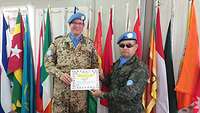 Ein VN-Soldat übergibt einem deutschen Soldaten eine Urkunde, im Hintergrund Flaggen 