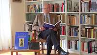 Ein Mann sitzt mit einem aufgeschlagenen Buch in den Händen vor einer Bücherwand.