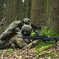 Zwei Soldaten gehen in einem Waldstück mit Waffe in Deckung.