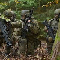 Bewaffnete Soldaten hocken kniend im Wald und blicken in eine Richtung.