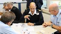 Zwei deutsche und ein französischer Soldat sitzen an einem Tisch. Der Franzose erläutert etwas und macht dazu eine Geste.
