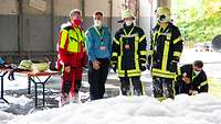 Fünf Männer, davon einer in Feuerwehruniform, vier in Schutzkleidung, nebeneinander im Schaum