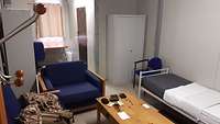 Ein Zimmer: Im Vordergrund ein Tisch mit Stühlen, daneben ein Bett und ein Schrank