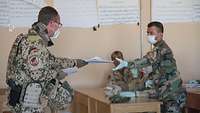 Ein deutscher Soldat überreicht einem afghanischen Soldaten in einem Unterrichtsraum einen Stapel Papier