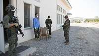 Ein bewaffneter finnischer Soldat vor einem Haus, im Hintergrund ziehen sich drei afghanische Soldaten Gummihandschuhe an 