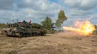 Ein Panzer beim Schießen im Gelände. Vor der Mündung der Kanone entsteht eine Feuerlanze.