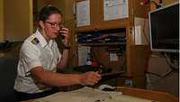 Eine schlanke, dunkelhaarige Frau mit Brille sitzt an einem Schreibtisch und telefoniert.