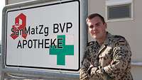 Ein deutscher Soldat lehnt mit verschränkten Armen an einem Schild mit der Aufschrift „Apotheke“, dahinter ein Containerhaus