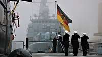 Marinesoldaten in dunkelblauer Uniform stehen auf einem Schiffsdeck, im Hintergrund hängt eine schwarz-rot-goldene Flagge.