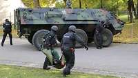 Zwei Polizeikräfte tragen einen Schwerverletzten auf einem Rettungstuch. Im Hintergrund steht ein Transportpanzer Fuchs