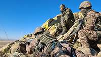 Ein Soldat liegt mit dem Gewehr im Anschlag hinter einem Felsen. Zwei weitere Soldaten beobachten den Schussverlauf.