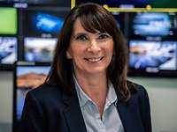 BAAINBw portrait handover studio A woman with dark hair, a dark blazer and a blouse stands smiling in front of a wall with monitors