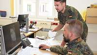 A soldier sitting at a computer. A further soldier looking over his shoulder.