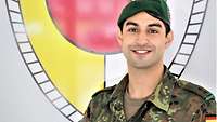Ein junger Soldat schaut lächelnd in die Kamera, hinter ihm das Verbandswappen.