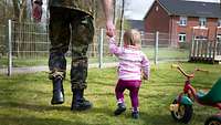 Ein Soldat hält seine kleine Tochter an der Hand. Daneben steht ein Dreirad.