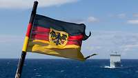 Die Dienstflagge der Marine weht im Wind am Heck eines Schiffes, im Hintergrund die offene See und ein britisches Schiff