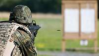 Ein Soldat in Flecktarn und Schießanzug steht mit einem Gewehr im Anschlag und schießt auf eine Schießwand.