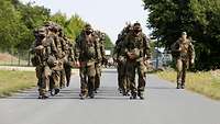 Eine Gruppe von Soldaten in Flecktarn und Gepäck maschieren in drei Reihen nebeneinander, ein Ausbilder geht rechts daneben.