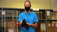 Ein Rekrut mit blauem T-Shirt und einer Mund-Nasenschutz-Maske probiert Handschuhe in Flecktarn an.