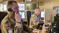 Zwei Soldaten sitze am Schreibtisch vor ihren Computern. Ein Soldat hält einen Telefonhöhrer a seinem Ohr.