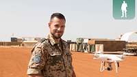 Ein Soldat steuert mit einer Fernbedienung eine Drohne vor sich, im Hintergrund mehrere Container 