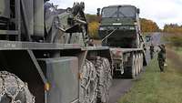 Ein Lastwagen zieht mit der Winde einen anderen Lastwagen auf die Ladefläche. Unscharf im Hintergrund ungarische Soldaten.