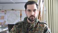 Ein Soldat im Feldanzug in einem Raum. Er blickt direkt in die Kamera. Im Hintergrund Lagekarten.