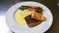 Ein Teller mit Lachsforellenfilet, Zitronenrisotto, Sauce Hollandaise und grünem Spargel im Speckmantel